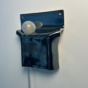 Peut inclure: Une applique murale en c&eacute;ramique bleu marine fonc&eacute; brillant avec une plaque arri&egrave;re rectangulaire dot&eacute;e de deux trous de fixation. Le luminaire pr&eacute;sente une section avant incurv&eacute;e en forme de poche contenant une ampoule blanche givr&eacute;e.
