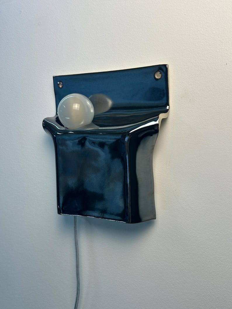 Peut inclure: Une applique murale en c&eacute;ramique bleu marine fonc&eacute; brillant avec une plaque arri&egrave;re rectangulaire dot&eacute;e de deux trous de fixation. Le luminaire pr&eacute;sente une section avant incurv&eacute;e en forme de poche contenant une ampoule blanche givr&eacute;e.