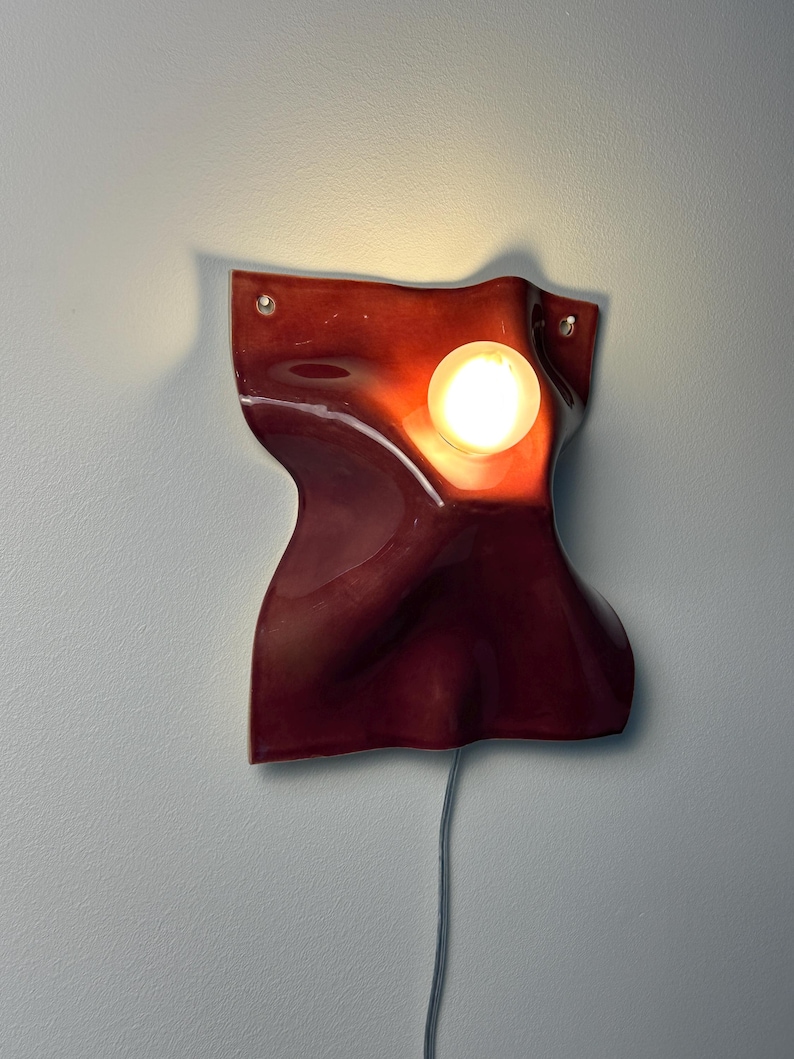 Puede incluir: Una l&aacute;mpara de pared con una pantalla de cer&aacute;mica de color rojo oscuro brillante, de forma org&aacute;nica y superficie ondulada. Una bombilla redonda e iluminada est&aacute; incrustada en la parte superior derecha. Un cable gris cuelga de la parte inferior.