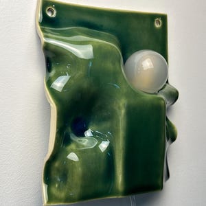 May include: A wall-mounted ceramic light fixture featuring a glossy, deep green, undulating surface with a sculptural, wave-like form. A single, spherical, frosted white glass light bulb is integrated into the right side of the green ceramic panel.