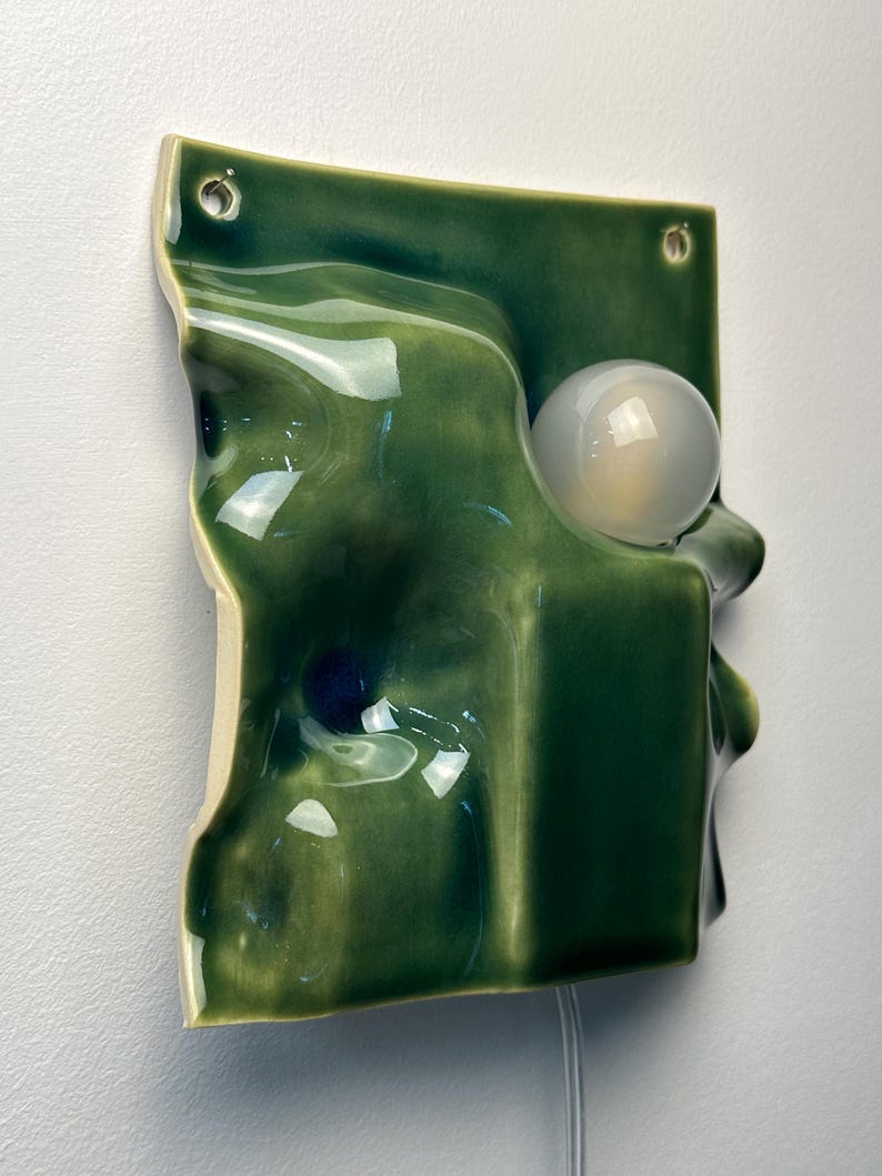 May include: A wall-mounted ceramic light fixture featuring a glossy, deep green, undulating surface with a sculptural, wave-like form. A single, spherical, frosted white glass light bulb is integrated into the right side of the green ceramic panel.