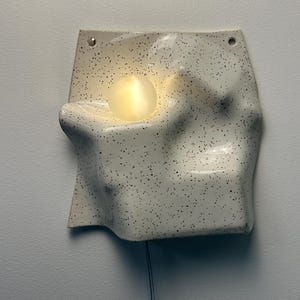 May include: A wall-mounted ceramic light fixture with an off-white, speckled finish and an irregular, undulating surface. A single, glowing, spherical light bulb is recessed into the center of the form, which features two small holes at the top.
