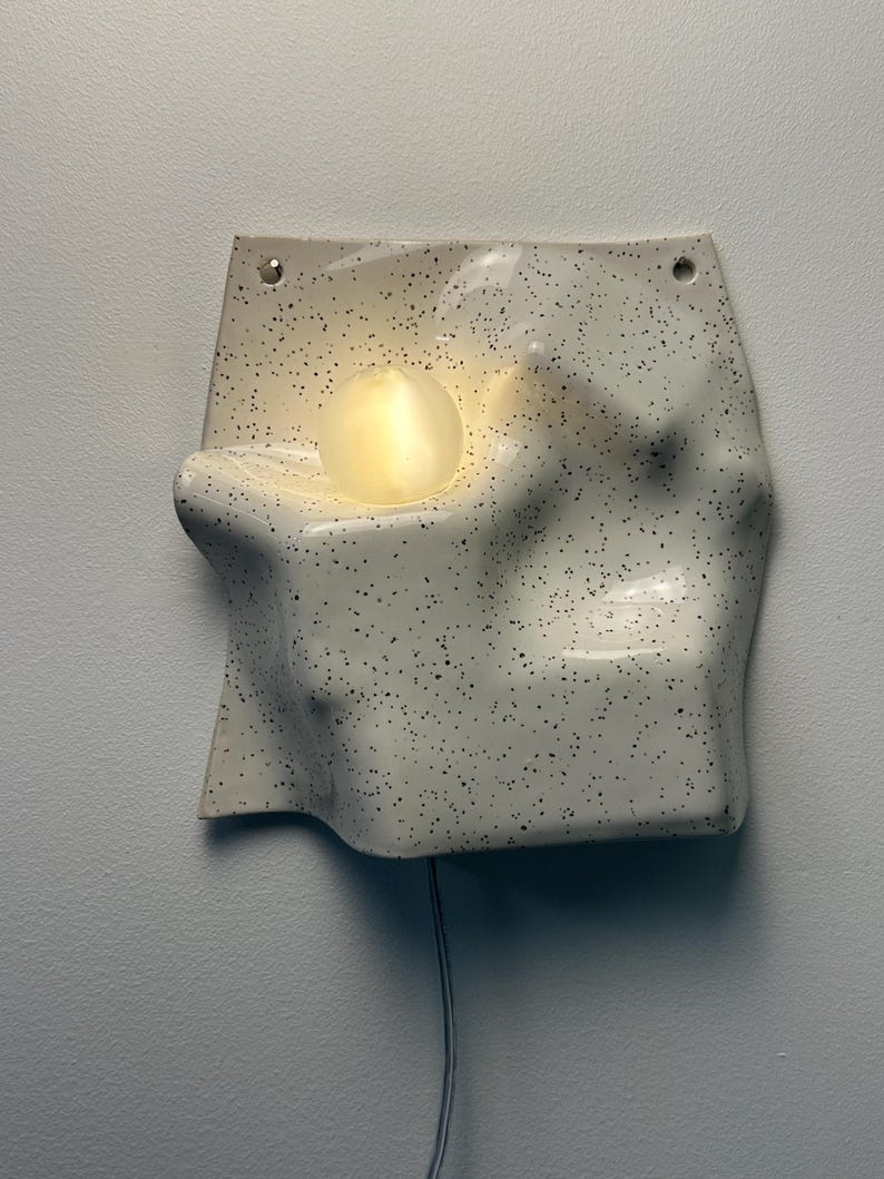 May include: A wall-mounted ceramic light fixture with an off-white, speckled finish and an irregular, undulating surface. A single, glowing, spherical light bulb is recessed into the center of the form, which features two small holes at the top.