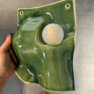 May include: A wall-mounted ceramic light fixture with a glossy, deep green glaze and an undulating, organic shape. The piece features two small circular holes at the top for mounting and a central, spherical white glass light bulb.