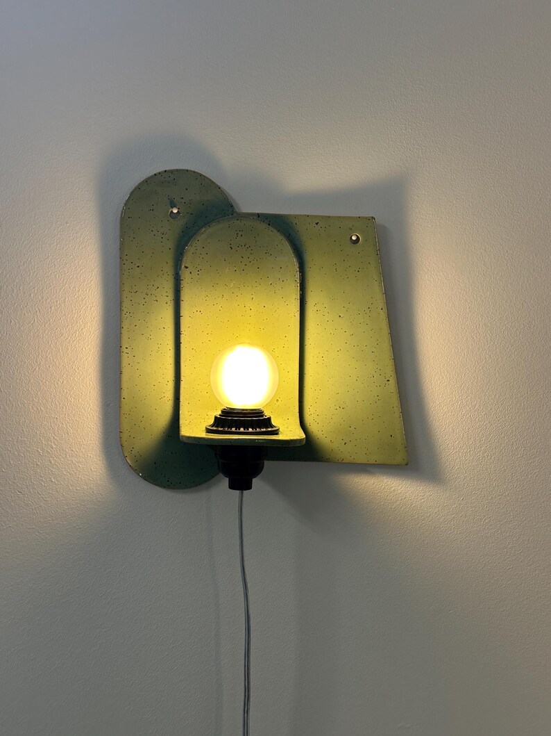 May include: A wall-mounted light fixture featuring a sage green speckled backplate with an arched center section. A single round, glowing light bulb is seated in a black socket attached to a small shelf, with a thin power cord extending downward.