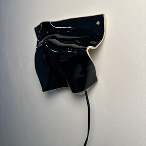 Puede incluir: Un aplique de pared con una pantalla de cer&aacute;mica negra brillante, de forma irregular y plegada, con un borde claro. Un fino cable de alimentaci&oacute;n negro sale de la parte inferior del objeto, montado contra una pared blanca lisa.
