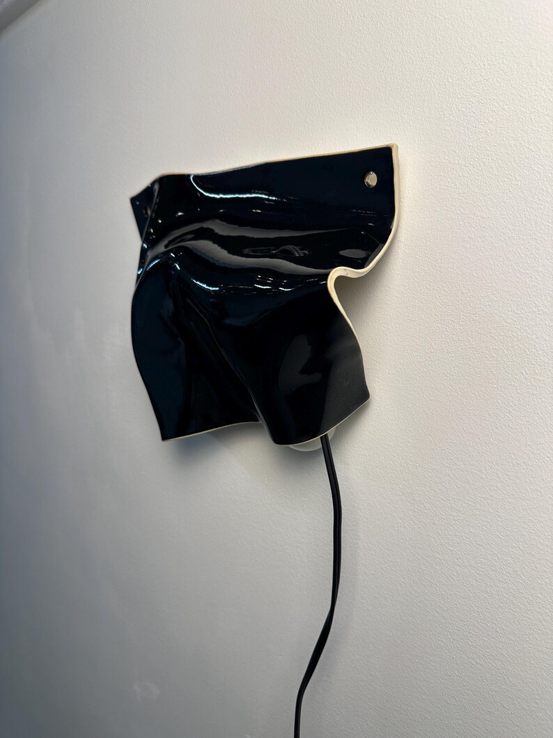 Puede incluir: Un aplique de pared con una pantalla de cer&aacute;mica negra brillante, de forma irregular y plegada, con un borde claro. Un fino cable de alimentaci&oacute;n negro sale de la parte inferior del objeto, montado contra una pared blanca lisa.