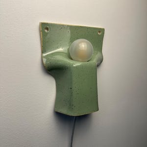 May include: A wall-mounted light fitting featuring a sage green ceramic base with a speckled finish. The fitting has a rectangular top section with two mounting holes and a curved, protruding lower section that holds a single round frosted light bulb.