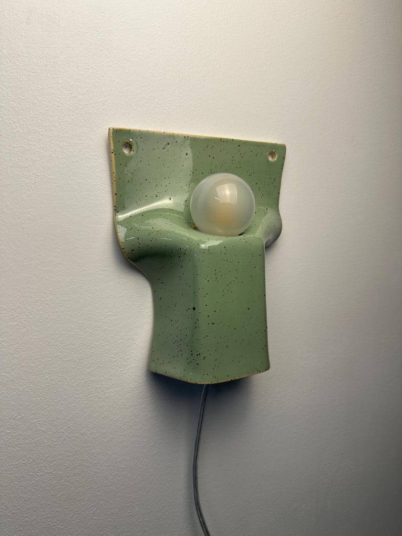 May include: A wall-mounted light fitting featuring a sage green ceramic base with a speckled finish. The fitting has a rectangular top section with two mounting holes and a curved, protruding lower section that holds a single round frosted light bulb.
