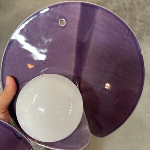 K&ouml;nnte beinhalten: Eine Wandleuchte mit einem gro&szlig;en, kreisf&ouml;rmigen, gl&auml;nzend violetten Keramikschirm mit einem einzelnen Befestigungsloch an der Oberseite. Eine kugelf&ouml;rmige, undurchsichtige wei&szlig;e Glaskugel ist am unteren Teil der Keramikscheibe befestigt.