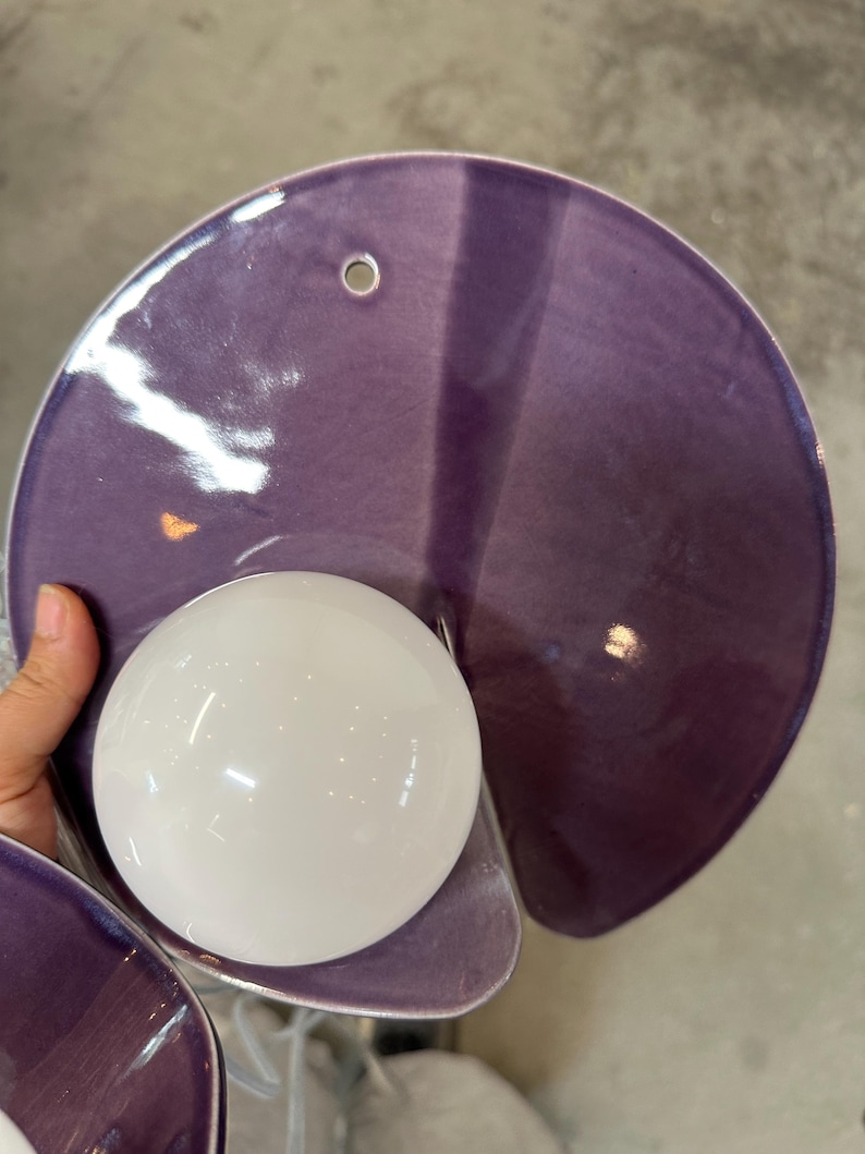 K&ouml;nnte beinhalten: Eine Wandleuchte mit einem gro&szlig;en, kreisf&ouml;rmigen, gl&auml;nzend violetten Keramikschirm mit einem einzelnen Befestigungsloch an der Oberseite. Eine kugelf&ouml;rmige, undurchsichtige wei&szlig;e Glaskugel ist am unteren Teil der Keramikscheibe befestigt.