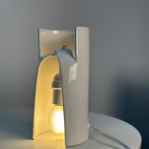 May include: A white ceramic table lamp with a curved, cylindrical structure that features an open front. A single light bulb is suspended inside the hollow center, emitting a warm glow. A thin, clear electrical cord extends from the base.