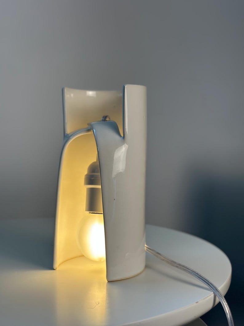 May include: A white ceramic table lamp with a curved, cylindrical structure that features an open front. A single light bulb is suspended inside the hollow center, emitting a warm glow. A thin, clear electrical cord extends from the base.