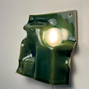 May include: A wall-mounted lamp featuring a glossy, dark green ceramic base with an irregular, undulating surface. The ceramic piece has two circular holes at the top corners and holds a single, round, frosted white glass light bulb.