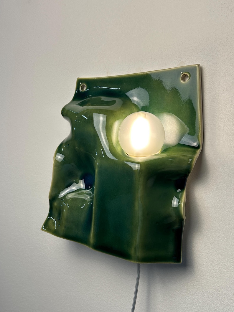 May include: A wall-mounted lamp featuring a glossy, dark green ceramic base with an irregular, undulating surface. The ceramic piece has two circular holes at the top corners and holds a single, round, frosted white glass light bulb.