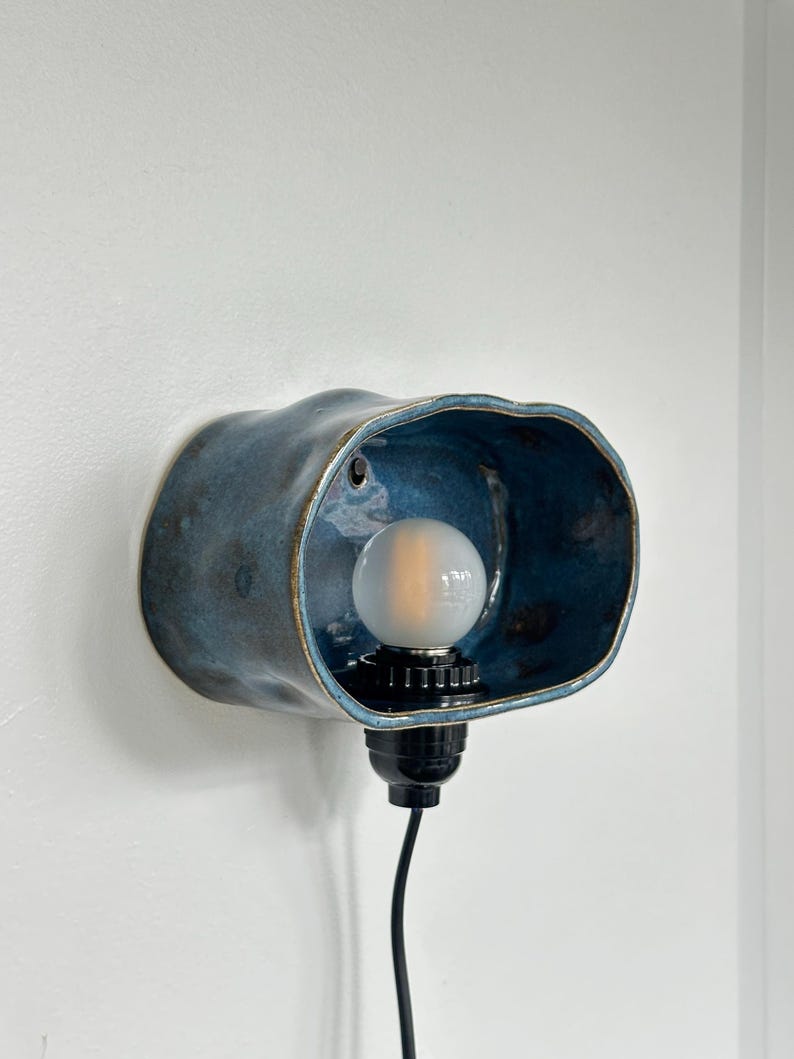 May include: A blue ceramic wall sconce with a bulb. The sconce has a unique, organic shape and a glossy finish. The interior of the sconce is a darker shade of blue. The bulb is white and round, and the fixture has a black base and cord.