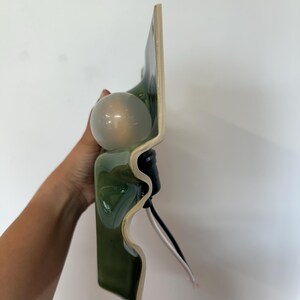 May include: A wall-mounted light fixture featuring a wavy, glossy dark green ceramic base with a smooth, curved profile. A single frosted white spherical light bulb is attached to the center, accompanied by a black socket and exposed electrical wires.