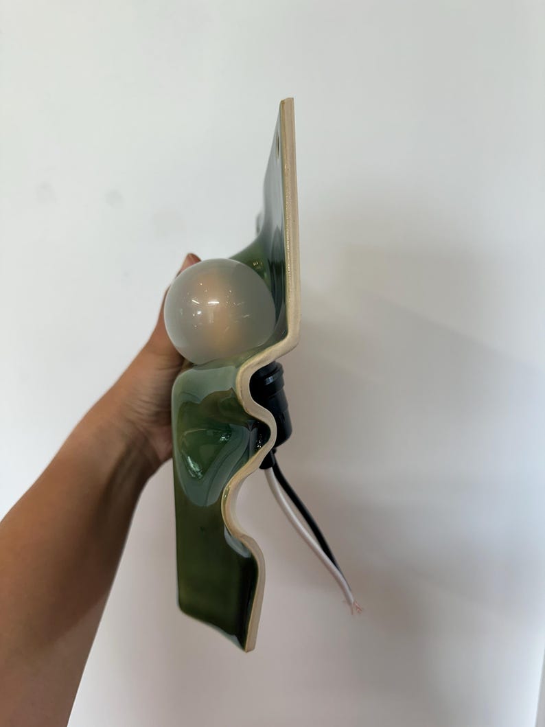 May include: A wall-mounted light fixture featuring a wavy, glossy dark green ceramic base with a smooth, curved profile. A single frosted white spherical light bulb is attached to the center, accompanied by a black socket and exposed electrical wires.