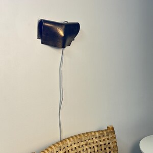May include: A dark brown, curved wall-mounted lamp with a smooth, metallic finish. The fixture features a folded, sculptural design and is connected to a thin, light-coloured electrical cord that hangs vertically downward from the base.
