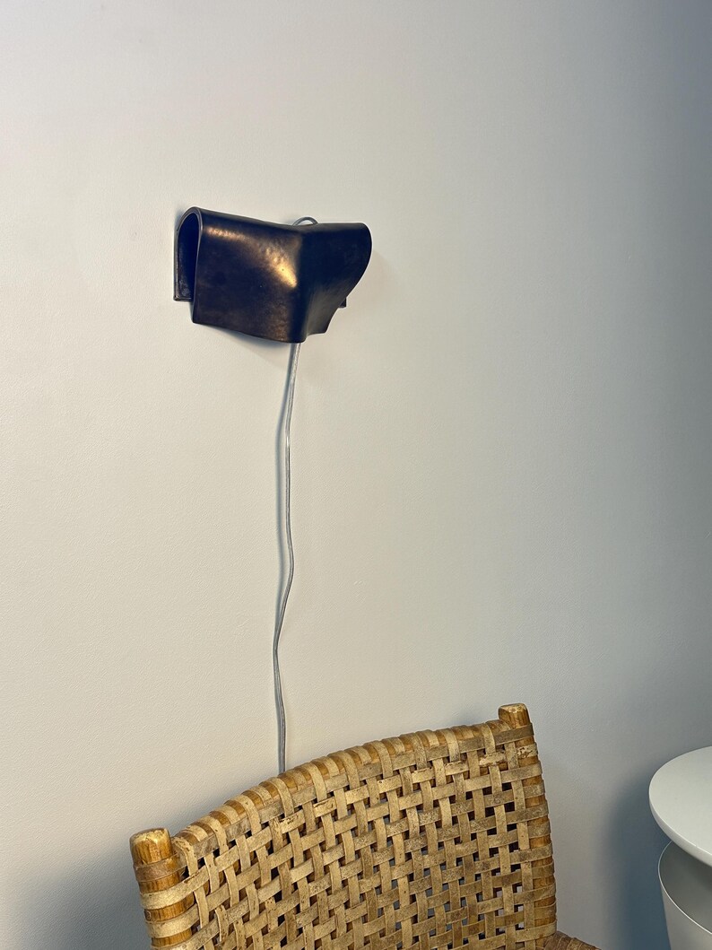 May include: A dark brown, curved wall-mounted lamp with a smooth, metallic finish. The fixture features a folded, sculptural design and is connected to a thin, light-coloured electrical cord that hangs vertically downward from the base.