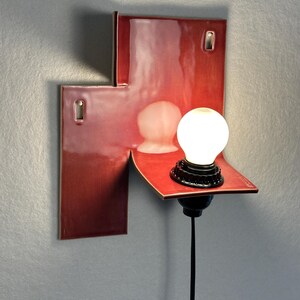 Pode incluir: Uma arandela de parede moderna com acabamento vermelho brilhante. A lumin&aacute;ria apresenta um design geom&eacute;trico com uma pequena prateleira que suporta um elemento de vidro fosco e uma l&acirc;mpada redonda e iluminada. Um cabo preto se estende para baixo.