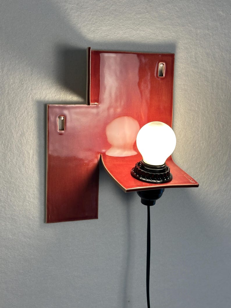 Pode incluir: Uma arandela de parede moderna com acabamento vermelho brilhante. A lumin&aacute;ria apresenta um design geom&eacute;trico com uma pequena prateleira que suporta um elemento de vidro fosco e uma l&acirc;mpada redonda e iluminada. Um cabo preto se estende para baixo.