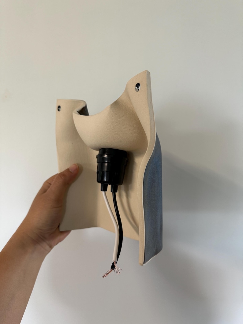 May include: A wall-mounted light fixture featuring a curved, matte beige ceramic shade with a dark blue exterior finish. The fixture includes a central black socket housing with two exposed electrical wires, one black and one white, extending downward.
