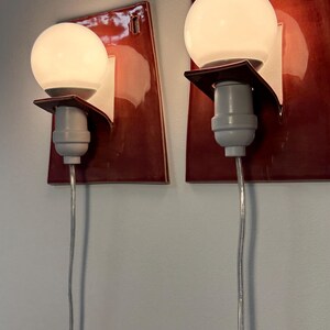Puede incluir: Un par de apliques de pared con placas traseras rectangulares de cer&aacute;mica de color rojo oscuro brillante. Cada l&aacute;mpara incluye una bombilla de cristal esf&eacute;rica blanca sujeta por un soporte blanco y un casquillo gris claro, con un cable transparente.
