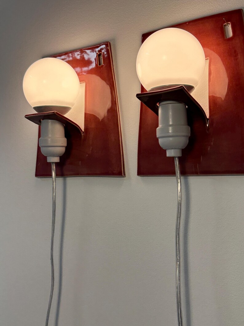 Puede incluir: Un par de apliques de pared con placas traseras rectangulares de cer&aacute;mica de color rojo oscuro brillante. Cada l&aacute;mpara incluye una bombilla de cristal esf&eacute;rica blanca sujeta por un soporte blanco y un casquillo gris claro, con un cable transparente.