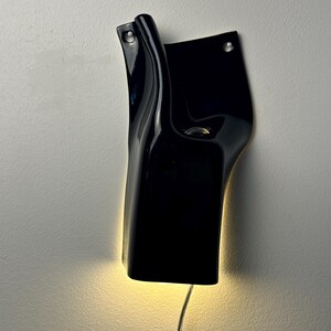 May include: A black wall-mounted light fixture with a glossy, curved, and asymmetrical form. The fixture features two circular mounting holes at the top and emits a warm, soft glow from its base, with a thin power cord extending downward.