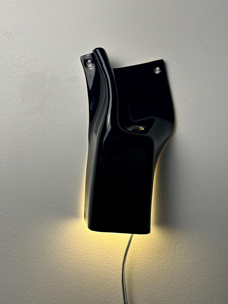 May include: A black wall-mounted light fixture with a glossy, curved, and asymmetrical form. The fixture features two circular mounting holes at the top and emits a warm, soft glow from its base, with a thin power cord extending downward.