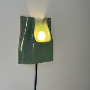 May include: A sage green ceramic wall sconce with a glossy finish and a curved, organic shape. The fixture features a central cutout housing a glowing, warm-toned spherical light bulb. A thin black power cord extends from the bottom of the unit.