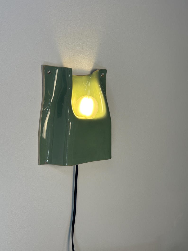 May include: A sage green ceramic wall sconce with a glossy finish and a curved, organic shape. The fixture features a central cutout housing a glowing, warm-toned spherical light bulb. A thin black power cord extends from the bottom of the unit.