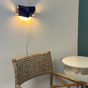 May include: A dark blue wall-mounted lamp with a curved, folded design emitting a warm glow from the top and bottom. Below the lamp, a woven rattan chair with a light brown frame sits next to a small, white, round tiered side table.