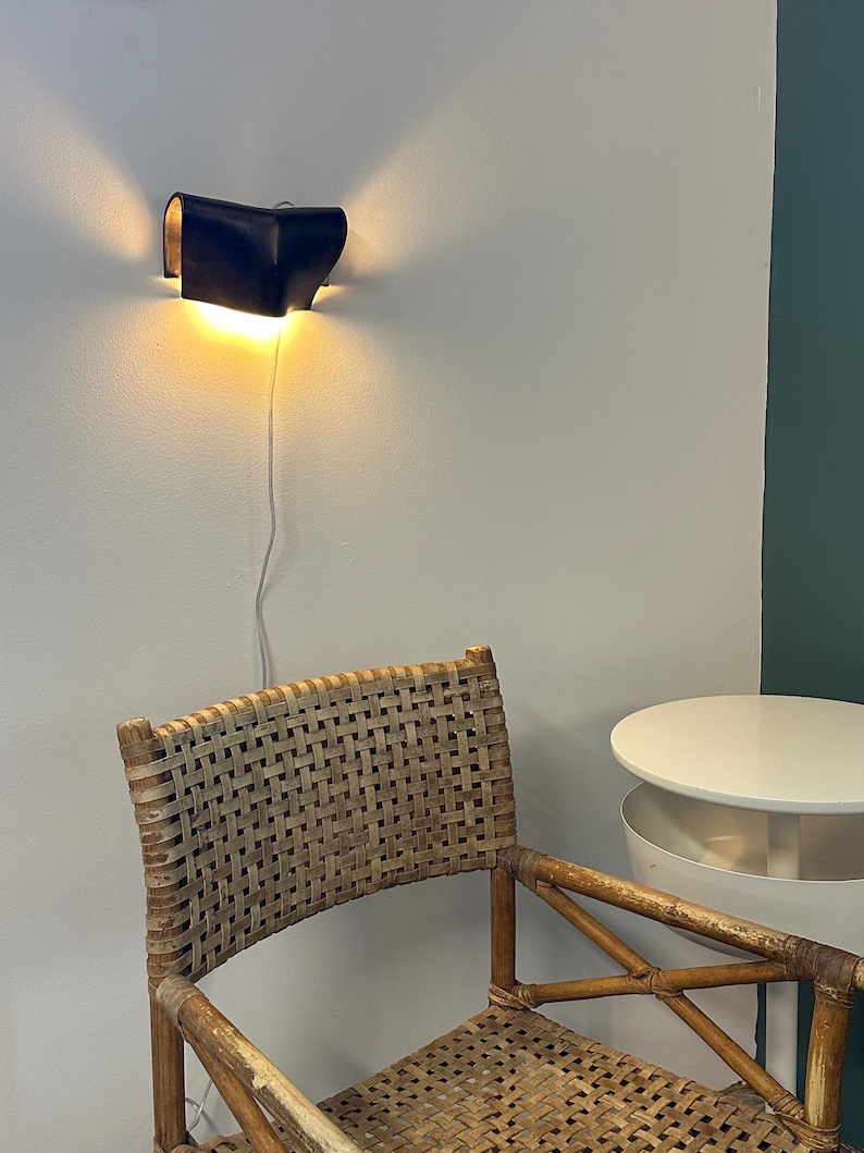 May include: A dark blue wall-mounted lamp with a curved, folded design emitting a warm glow from the top and bottom. Below the lamp, a woven rattan chair with a light brown frame sits next to a small, white, round tiered side table.
