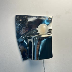 K&ouml;nnte beinhalten: Eine Wandleuchte mit einer reflektierenden, silberfarbenen Metallplatte in unregelm&auml;&szlig;iger, geschwungener Form. Die Platte weist auf der linken Seite ein Raster aus kreisf&ouml;rmigen Vertiefungen auf und ist mit einer mattwei&szlig;en Kugellampe versehen.