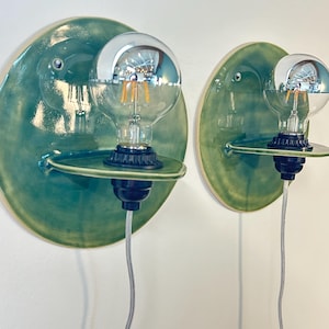 May include: Two wall-mounted light fixtures featuring circular, glossy green ceramic backplates. Each fixture has a small, horizontal ceramic shelf extending from the center, holding a black socket with a clear, round glass light bulb.