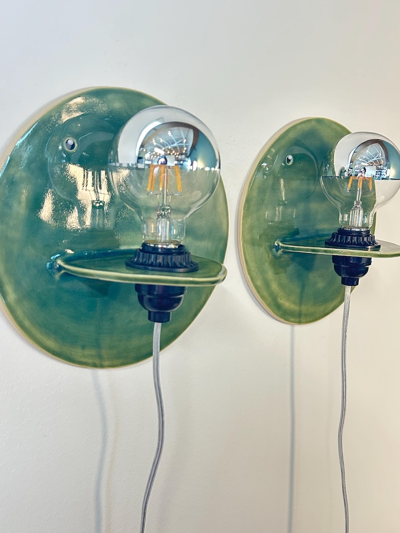 May include: Two wall-mounted light fixtures featuring circular, glossy green ceramic backplates. Each fixture has a small, horizontal ceramic shelf extending from the center, holding a black socket with a clear, round glass light bulb.