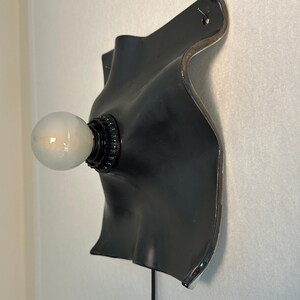 May include: A wall-mounted lamp featuring a matte black, curved, organic-shaped ceramic backplate with a glossy silver edge. A black socket holds a single, round, frosted white light bulb that extends outward from the center of the fixture.