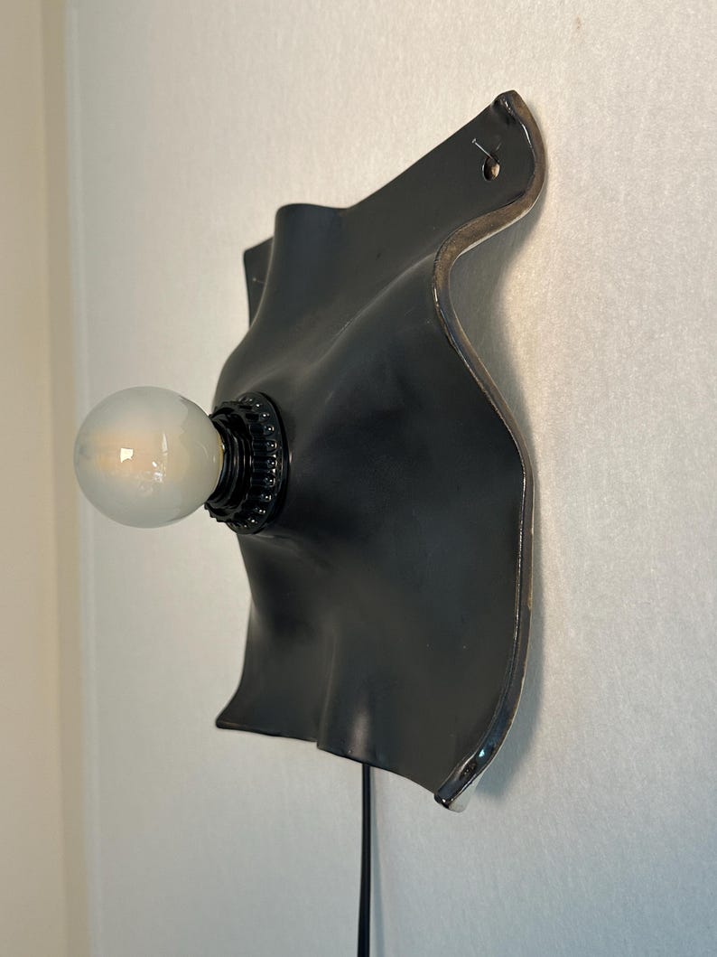 May include: A wall-mounted lamp featuring a matte black, curved, organic-shaped ceramic backplate with a glossy silver edge. A black socket holds a single, round, frosted white light bulb that extends outward from the center of the fixture.