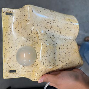 May include: A wall-mounted light fixture with a wavy, cream-colored ceramic surface featuring dark brown speckles. The fixture includes a single, round, frosted glass bulb integrated into the lower left section and two rectangular mounting slots.