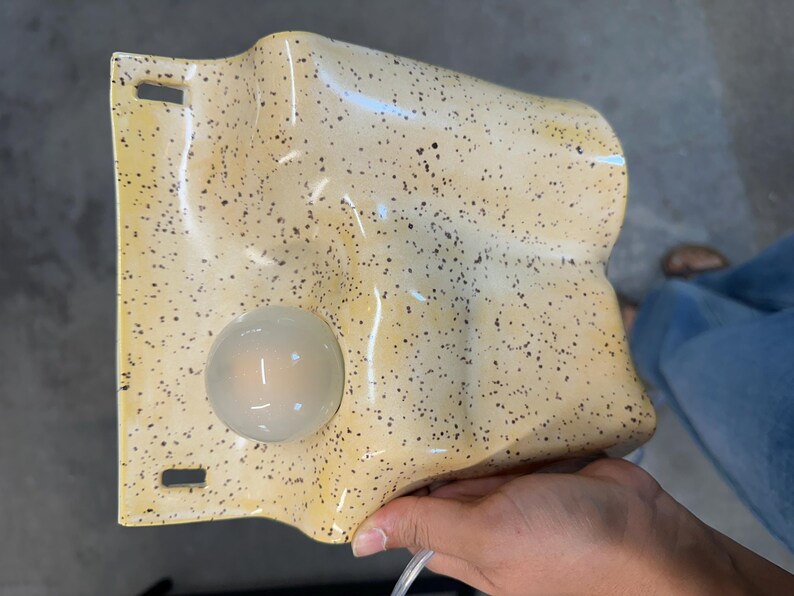 May include: A wall-mounted light fixture with a wavy, cream-colored ceramic surface featuring dark brown speckles. The fixture includes a single, round, frosted glass bulb integrated into the lower left section and two rectangular mounting slots.