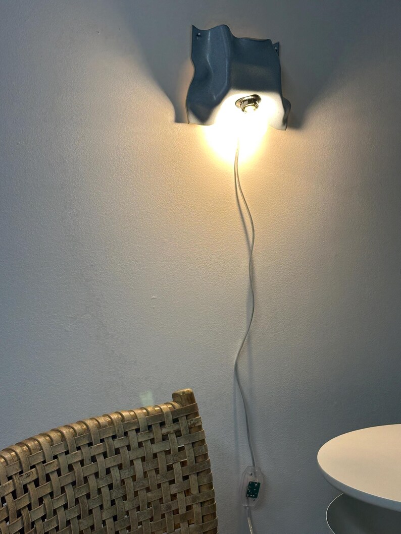 May include: A light blue wall-mounted lamp with a wavy, organic shape and a central light bulb emitting a warm glow. A thin white power cord extends downward from the fixture, connecting to a small clear switch near the bottom of the wall.