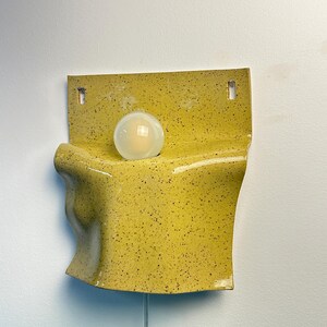 May include: A wall-mounted ceramic lamp featuring a bright yellow glaze with dark speckles. The fixture has a curved, organic shape with a flat top section containing two mounting holes and a single round, frosted white light bulb centered.