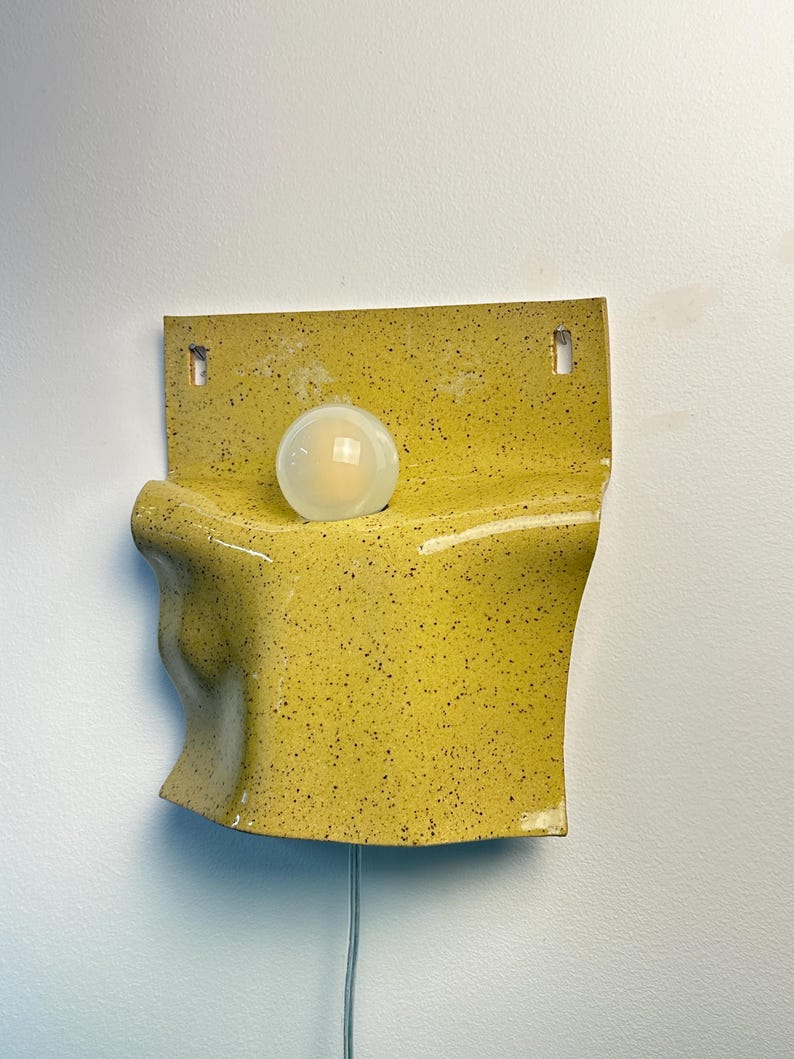 May include: A wall-mounted ceramic lamp featuring a bright yellow glaze with dark speckles. The fixture has a curved, organic shape with a flat top section containing two mounting holes and a single round, frosted white light bulb centered.