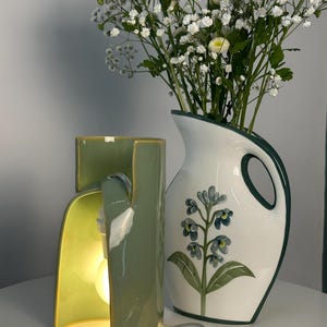 May include: A sage green ceramic table lamp with a curved, open-sided structure and an illuminated bulb, positioned next to a white ceramic pitcher featuring a dark green rim, a handle, and a painted blue floral design filled with white baby's breath.