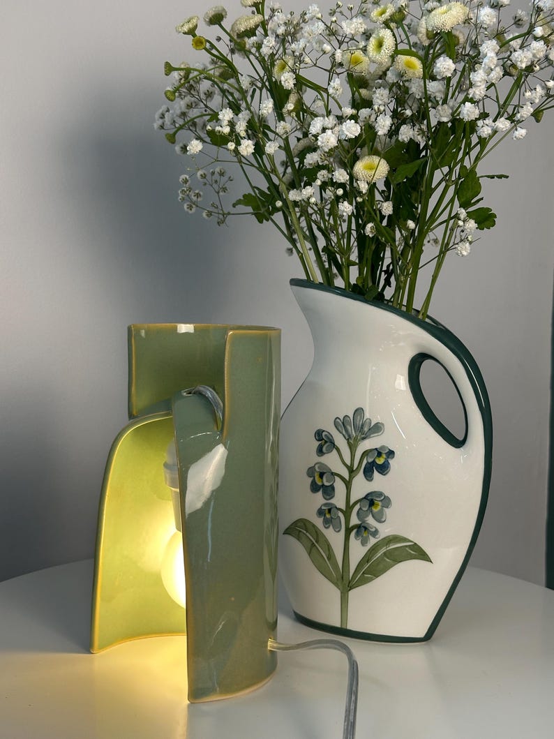 May include: A sage green ceramic table lamp with a curved, open-sided structure and an illuminated bulb, positioned next to a white ceramic pitcher featuring a dark green rim, a handle, and a painted blue floral design filled with white baby's breath.