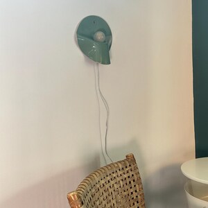 May include: A wall-mounted light fixture featuring a circular, sage green ceramic shade with a curved, downward-sloping design. A single white spherical light bulb is centered within the shade. A thin, white electrical cord hangs vertically downward.