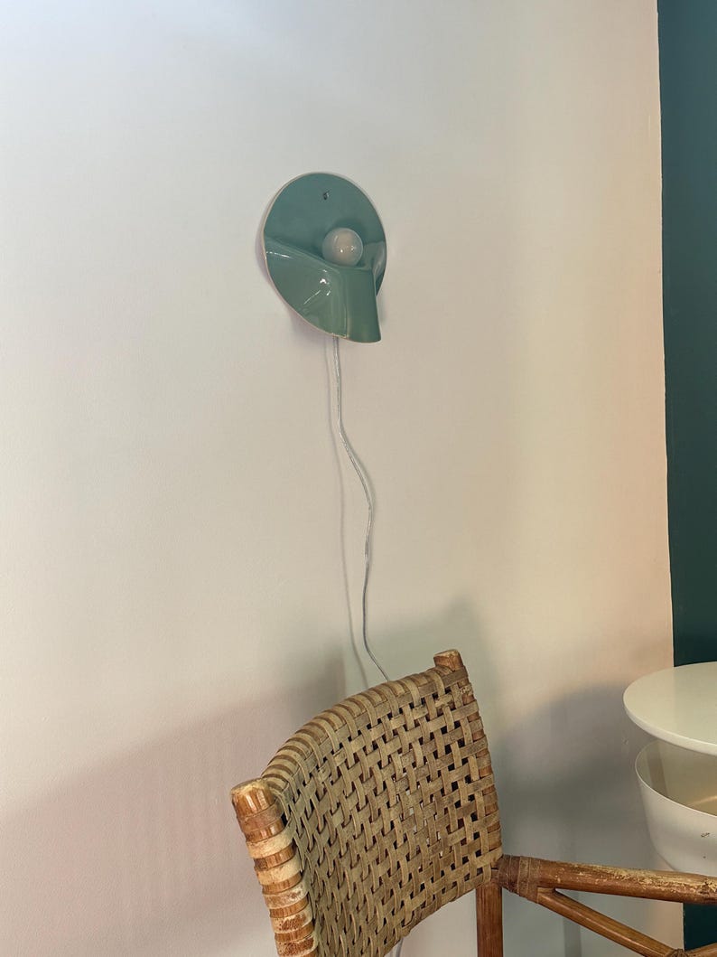 May include: A wall-mounted light fixture featuring a circular, sage green ceramic shade with a curved, downward-sloping design. A single white spherical light bulb is centered within the shade. A thin, white electrical cord hangs vertically downward.