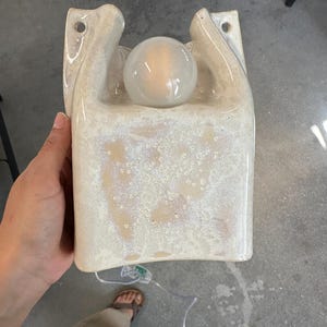 May include: A wall-mounted ceramic sconce with a glossy, cream-coloured speckled glaze. The fixture features a U-shaped cutout at the top, two circular mounting holes, and a single round, glowing light bulb centred within the ceramic housing.